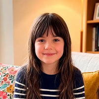 A young girl with long brown hair and bangs smiling, wearing a navy blue sweater with white horizontal stripes, sitting in a cozy indoor setting with a floral pillow and a bookshelf in the background.